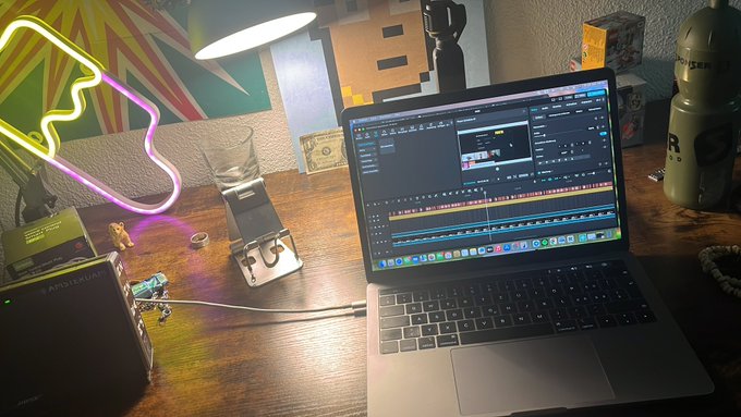 The image shows a laptop computer sitting on top of a wooden desk, with a neon sign behind it that reads "clipping clipping clipping da bois". The background features a colorful painting or poster.