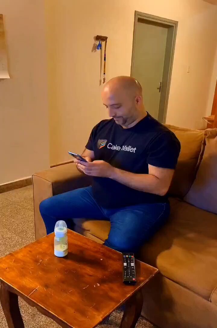 The image shows a man sitting on a couch with his legs crossed while looking at his cell phone. The man appears to be wearing a black shirt, and there are two remote controls placed next to him on the couch. A bottle of water is also present in front of him. The scene takes place in a living room setting, which suggests that they might have been watching a movie or television show together using one of the remotes.