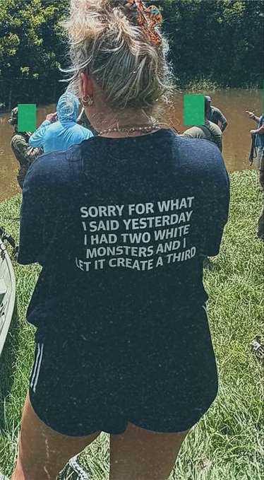 A woman with a bun in a hair scrunchie stands facing away from the camera at an outdoor gathering by a river, wearing a black t-shirt that reads "SORRY FOR WHAT I SAID YESTERDAY I HAD TWO WHITE MONSTERS AND I LET IT CREATE A THIRD."
