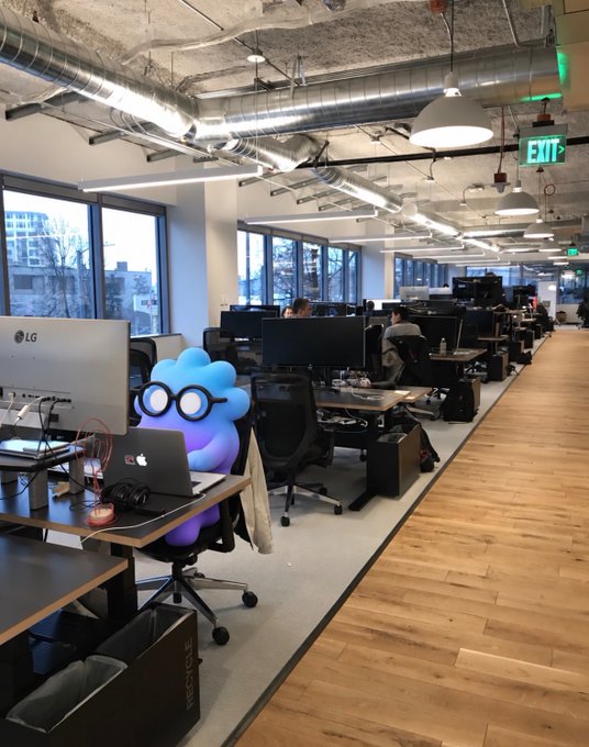 An open-plan office space with rows of desks and monitors, featuring a cute blue cloud character with round glasses sitting in an office chair at a workstation.