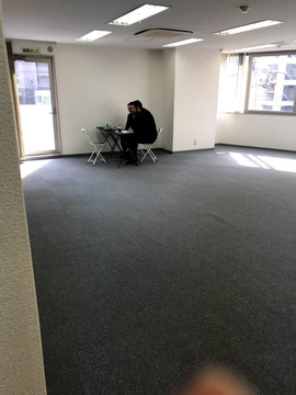 A man sits alone at a small table in a large, empty carpeted office space with white walls and fluorescent lighting, capturing the humble beginnings of Binance's original office in 2017 shortly after China's crypto crackdown. The "EXIT" sign is visible above a door on the left side of the room.