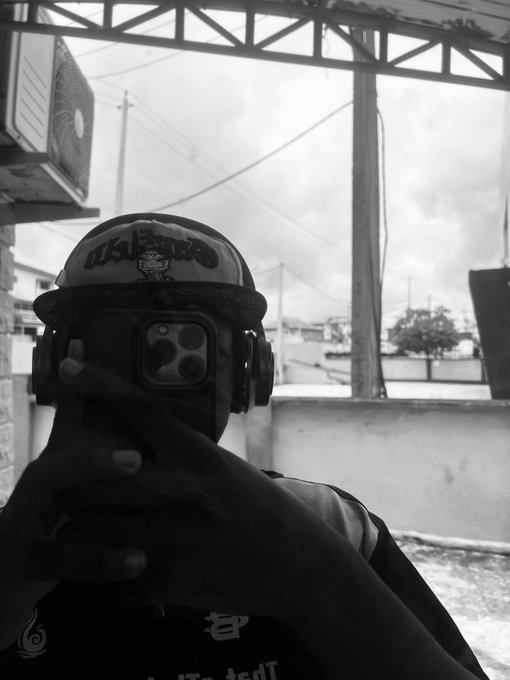 A black-and-white mirror selfie of a person wearing a 'BULLS' cap and large headphones, obscuring their face with a smartphone featuring a triple-camera setup.