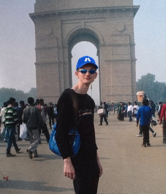 A young man wearing a blue cap, sunglasses, and a black shirt poses for a photo in front of the India Gate monument in New Delhi, with crowds of people walking around.