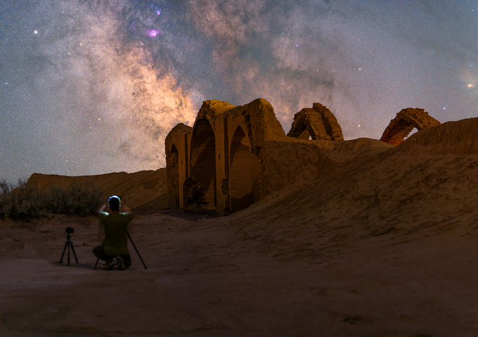 A night photograph of a photographer in a green chair photographing the Milky Way galaxy above ancient mud-brick ruins with arched structures in a desert landscape.