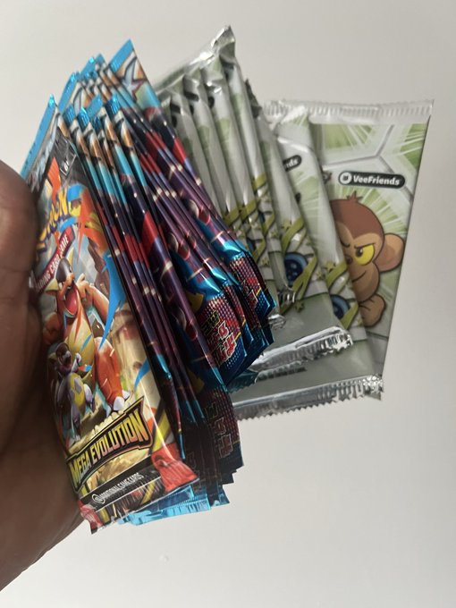 A hand holding a fan of unopened Pokémon trading card booster packs, including "MEGA EVOLUTION" and what appears to be a Japanese set, along with "VeeFriends" packs in the background.