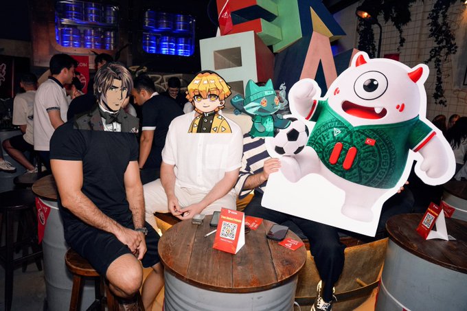 Three people sit at barrel tables in a dimly lit venue holding anime-style character cutouts of a soccer player and a white monster character, with a QR code menu on the table.