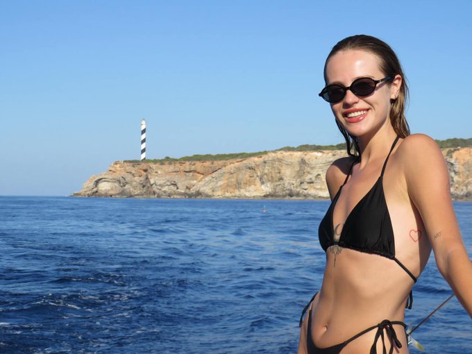 A young woman in a black bikini and sunglasses smiles at the camera on a boat, with a rocky coastline and a black-and-white striped lighthouse in the background.