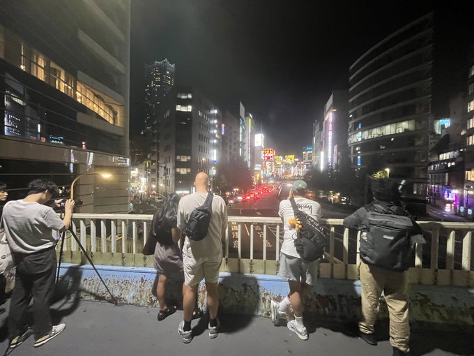 A group of photographers and onlookers stand on an elevated pedestrian walkway at night, observing a busy city street with neon signs and traffic below.