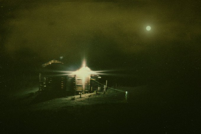 A dark, grainy night photograph showing a brightly lit building with a prominent starburst light effect and a full moon in the sky.