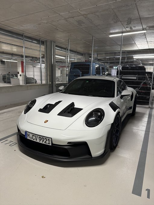 A white Porsche 911 GT3 with the German license plate 'M-CV 9921' is parked in a numbered spot inside a parking garage.