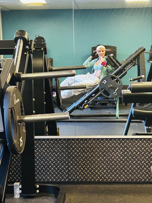 A gym scene showing exercise equipment in the foreground with a reflection in the mirror of a person wearing a blue outfit and head covering sitting on workout equipment.