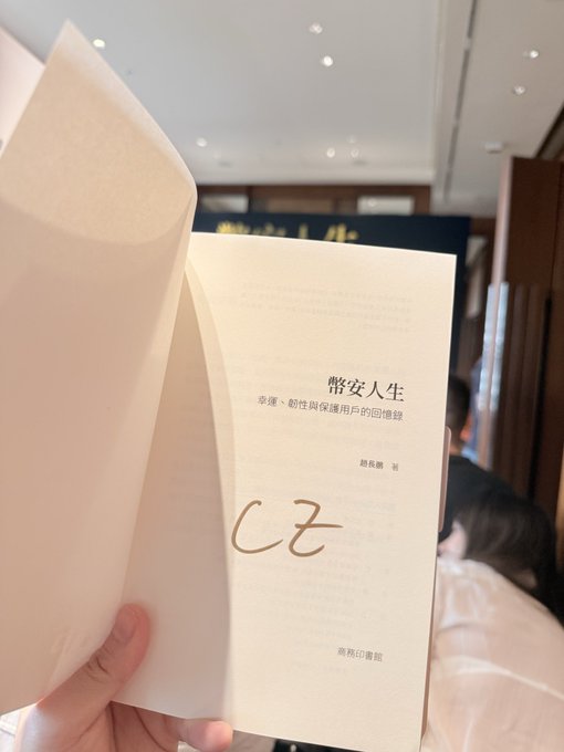 A hand holds open a book titled "幣安人生：掌握、韌性與保護用戶的回憶錄" (Binance Life: Mastery, Resilience, and Protecting User Memories) by CZ, revealing a stylized "CZ" signature on the title page. The image showcases the sought-after signed copy mentioned in the post, waiting for CZ (Changpeng Zhao, founder of Binance) to arrive and greet fans.