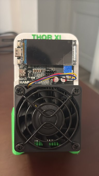 A homemade cryptocurrency mining rig labeled 'THOR XI' featuring a small circuit board with display and a large black cooling fan mounted on a green 3D-printed base.