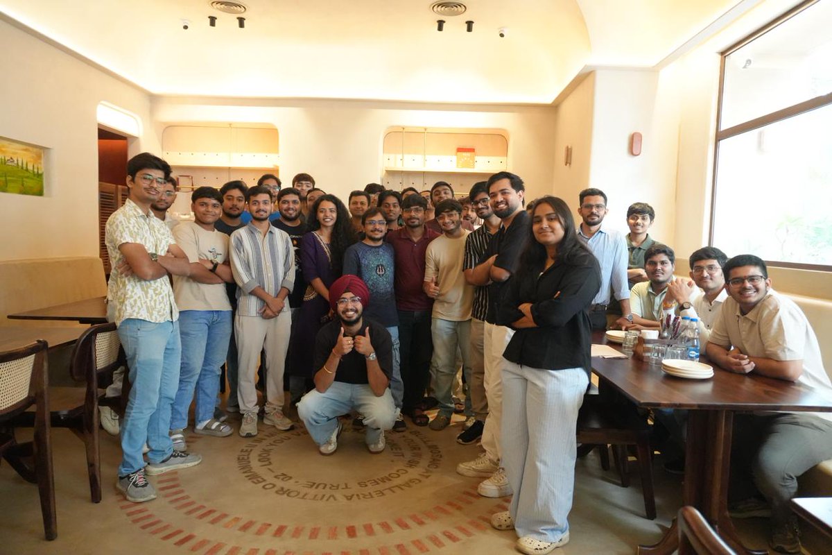 A large group of approximately 25 people poses together in what appears to be a restaurant or café, with one man kneeling in the center giving a thumbs-up gesture while surrounded by smiling attendees. The image captures attendees of the first ETH Gujarat gathering, with visible text on the circular floor rug reading "GALLERIA VITTORIO EMANUELE" alongside Italian lettering from the restaurant's flooring.