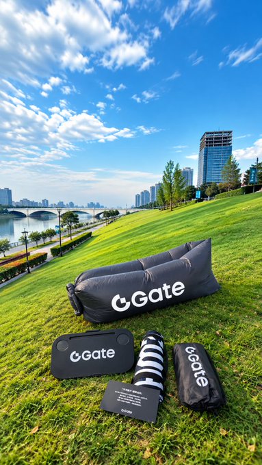 A set of Gate-branded camping accessories—including an inflatable sofa, picnic mat, and small table—laid out on a grassy riverside park, with the "Gate" logo clearly visible on multiple items. The scene perfectly complements the post about enjoying a peaceful outdoor picnic by the canal, showcasing the brand's stylish and practical giveaway gear.