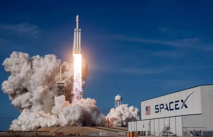 A SpaceX rocket launches from a pad beside a large hangar displaying the "SPACEX" logo and an American flag, with billowing clouds of smoke and bright exhaust trailing the ascending vehicle. The image symbolizes SpaceX's explosive growth as a company, mirroring the post's description of ticker "SPCX" (likely referring to a SpaceX-related stock) being targeted by massive short-selling bets worth over $25 billion.