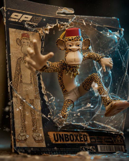 The image features a toy monkey figurine, which appears to be broken or damaged. It has leopard print shorts and a red hat that matches its outfit. The monkey figurine is placed inside an unopened package with a cracked window on top of it. The packaging also includes a label that reads "gm apes bored ape yacht club x.com/edizkan_". This suggests that the toy was recently purchased or received as a gift, and now it has been opened to reveal its contents.