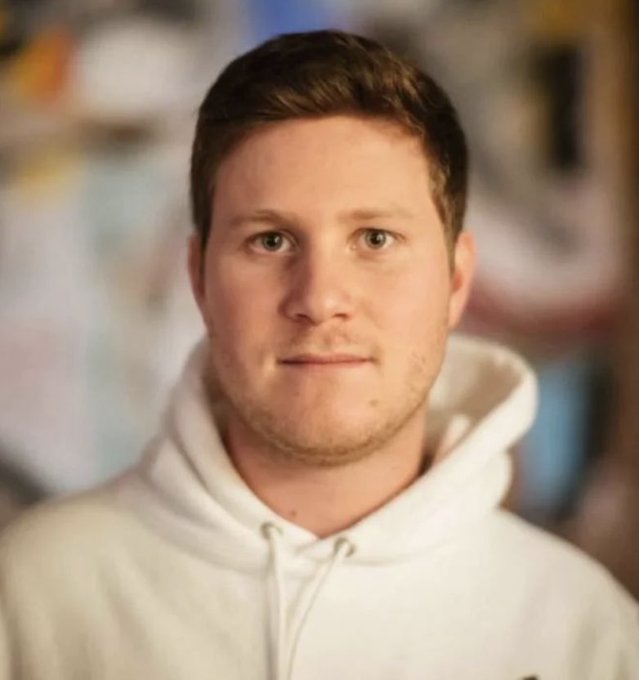 A headshot portrait of a young man with short brown hair and green eyes wearing a white Nike hoodie.