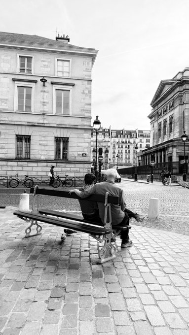 A black and white photo shows a couple sitting on a bench in a cobblestone plaza, looking at each other affectionately, surrounded by classic European architecture.