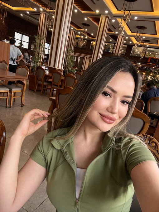 A selfie of a young woman with long highlighted hair wearing a green top, taken in a restaurant setting.