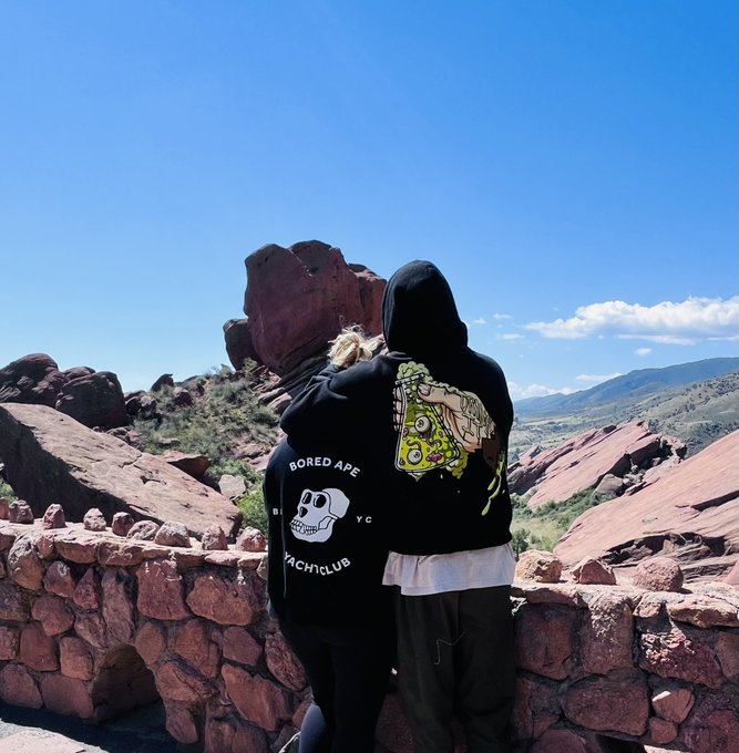 Two people wearing black Bored Ape Yacht Club hoodies (one with blonde hair visible) standing together overlooking a scenic landscape of red rock formations and distant mountains under a bright blue sky.