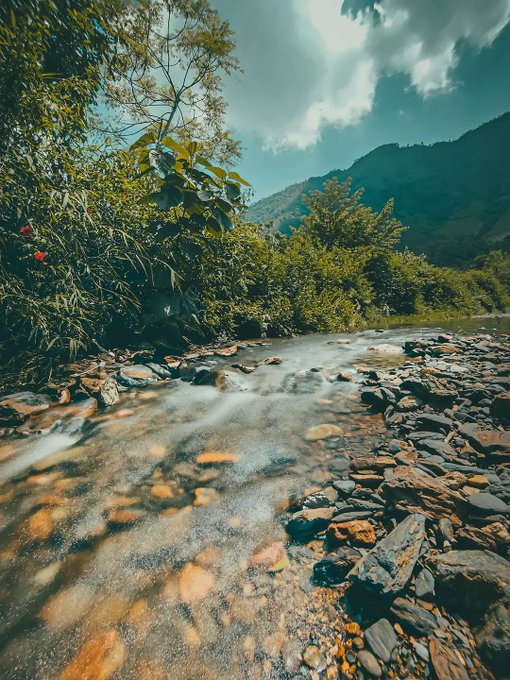 A scenic photograph of a rocky stream flowing through a lush green landscape with mountains and cloudy sky in the background.