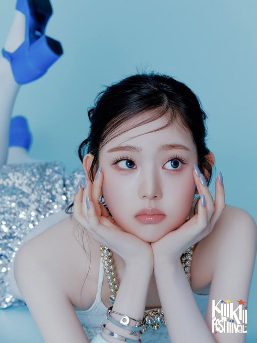 A young woman with long dark hair and soft makeup poses with her hands cupping her face, wearing a white top with beaded straps and silver bracelets. The image has a cool blue-toned aesthetic, with blue floral elements and a sparkly silver background, and features a "KiiiKiii Festival" logo in the bottom right corner.