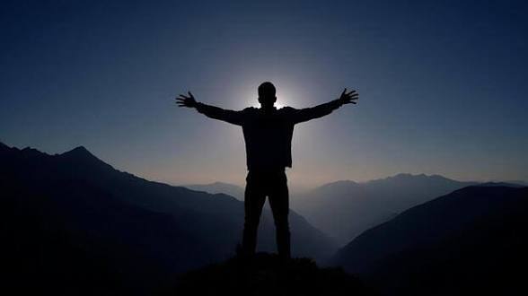 A silhouetted figure stands triumphantly with arms outstretched against a dramatic sunset over a mountainous landscape.