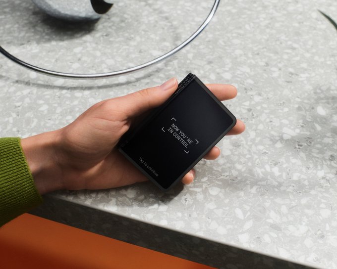 A hand holds a Ledger hardware wallet displaying "NOW YOU SEE I'M IN CONTROL" and "Tap to continue" on its screen, resting on a speckled countertop beside a cable. The device visually reinforces the post's message about the Ledger AI Security Roadmap, symbolizing hardware-based approval as a reliable safeguard against rogue AI agents operating without human awareness.