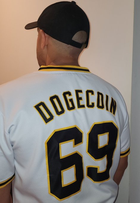 The image features a man wearing a white baseball uniform with black numbers, specifically the number 69, on his back. The background of the photo shows a wall, which could be part of a room or a hallway. There are no other people visible in the picture.