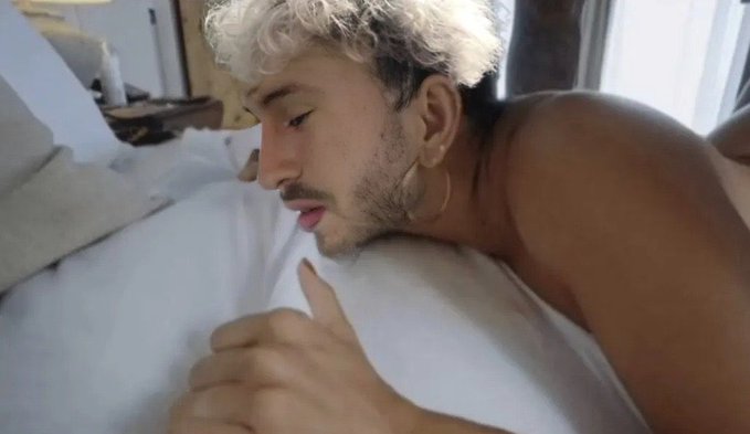 A shirtless man with bleached blonde curly hair and a beard lies in bed, looking down at his hand. He has a hoop earring and a small mark on his chest.