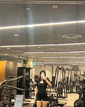 A woman in a black athletic outfit takes a mirror selfie inside a well-lit gym filled with various exercise equipment.