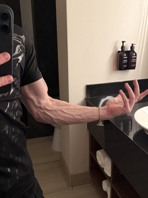 A muscular arm flexed in a bathroom mirror selfie, showcasing prominent veins in the bicep and forearm, perfectly capturing the "time to bulk" mentality with its emphasis on size and definition.