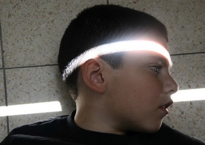A side-profile photo of a young person with short dark hair against a tiled wall with horizontal light streaks.