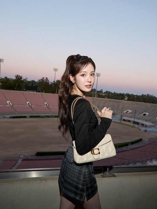 A young woman with long wavy brown hair stands in an empty stadium at sunset, wearing a black long-sleeve top, plaid skirt, and carrying a white quilted Gucci handbag.
