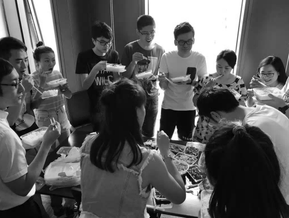 A black-and-white photo captures a group of coworkers gathered around a table sharing a meal, likely representing the early Binance team that received the iconic 20,000 BNB tokens (now worth $15 million) during the 2017 ICO. The casual office celebration scene reflects the humble beginnings of the company before its meteoric rise.