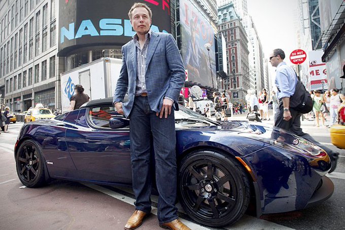 A man in a blue plaid jacket and jeans stands in front of a dark blue sports car on a busy city street, with a large digital billboard in the background.