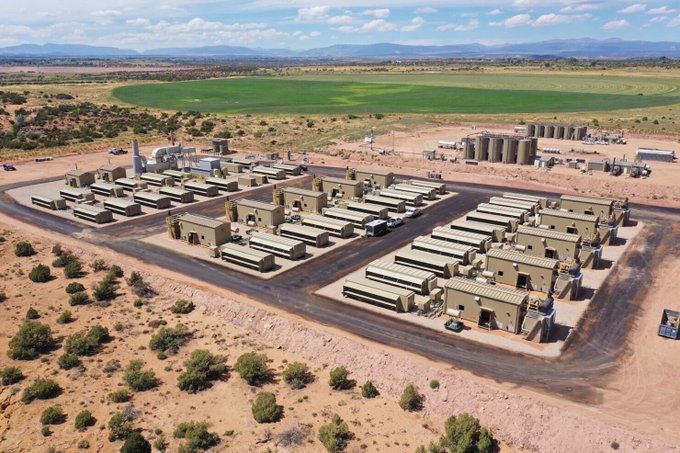 An aerial view of a Crusoe Energy data center facility, showing rows of shipping-container-style modular buildings housing Bitcoin mining equipment, alongside silos and infrastructure at what appears to be an oil field site in North Dakota. The image visually represents the company's pioneering approach of using flared natural gas from oil wells to power cryptocurrency mining operations.