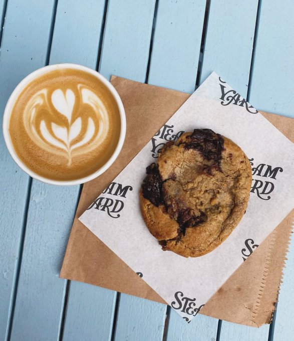 A latte with a tulip-shaped latte art design sits beside a large chocolate chunk cookie on a branded paper bag, both resting on a light blue wooden table.