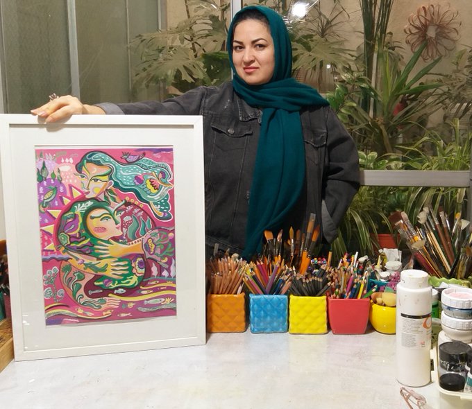 An artist in a teal headscarf and denim jacket poses next to her framed colorful illustration and a table filled with art supplies.