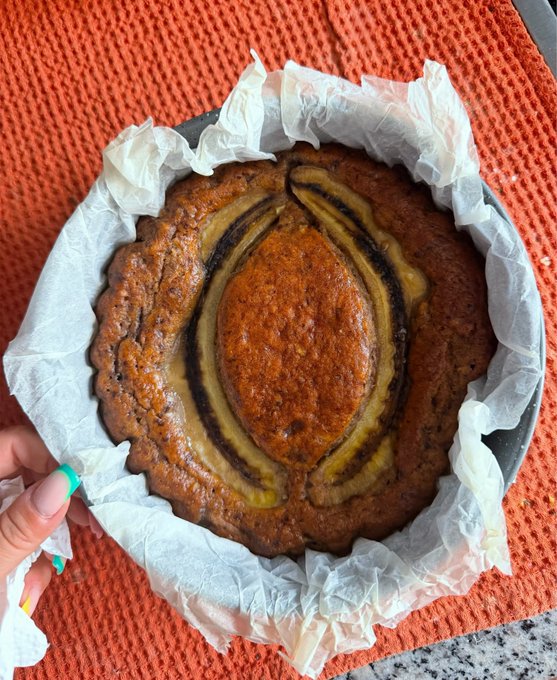 A freshly baked cake with a swirled peanut butter and jelly/jam pattern in a parchment-lined metal pan, held by a hand with green nail polish over an orange textured towel.