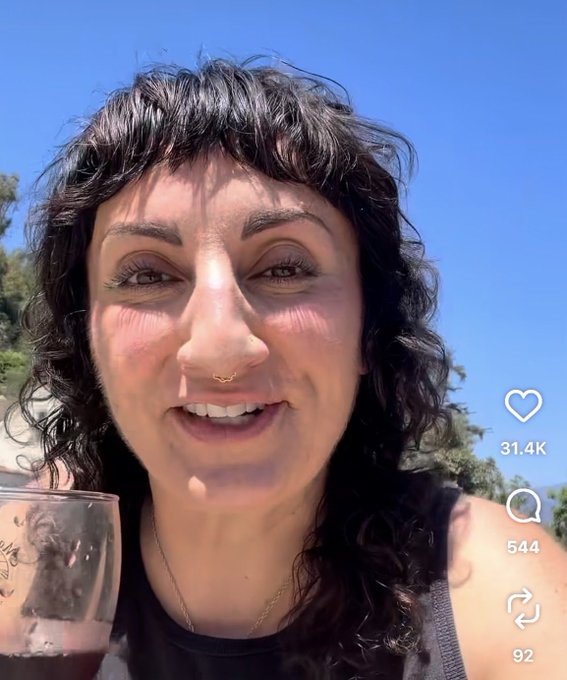 A selfie of a smiling woman with dark curly hair and a nose ring, holding a glass of red wine outdoors, with social media engagement icons showing 31.4K likes, 544 comments, and 92 shares.