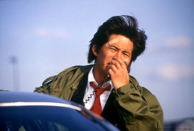 The caption should describe a man with dark, windswept hair wearing an olive green jacket, white shirt, and red tie, with a bandaged forehead and sunglasses hanging from his collar, appearing to be in distress or contemplation against a cloudy sky.

The man’s pained expression and bandaged forehead visually echo the post’s sentiment of "仮想通貨止まりません！！" (Cryptocurrency won't stop [falling]!!), humorously embodying the suffering of investors watching their crypto portfolios plummet.
