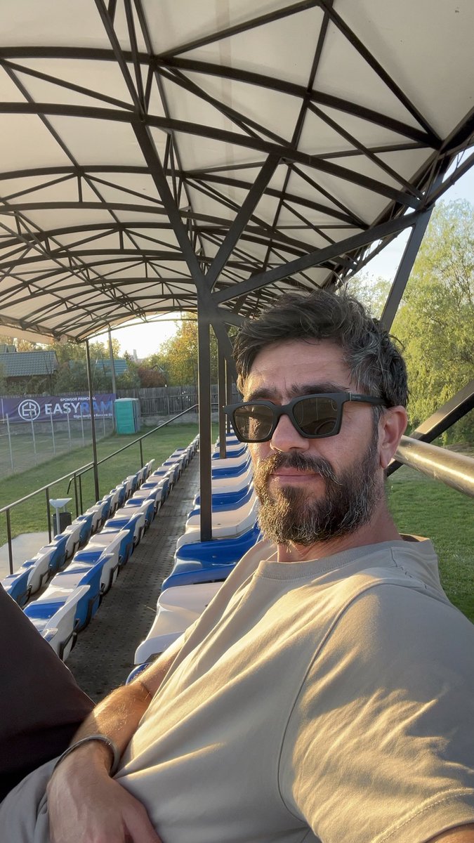 A bearded man wearing sunglasses sits under a metal canopy at a stadium with empty blue and white seats, with an 'EASYFAM' banner visible in the background.
