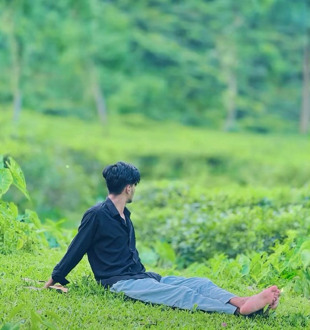A young man in a black shirt and gray pants sits barefoot in a lush green tea plantation, looking out over the verdant landscape.