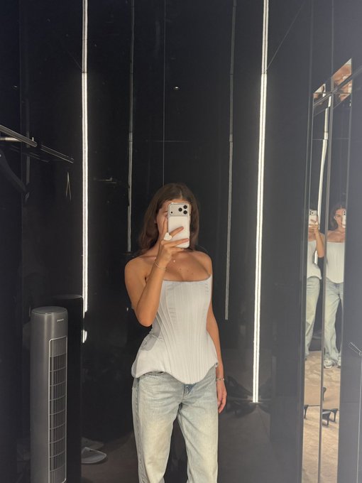 A mirror selfie of a woman with shoulder-length hair wearing a white strapless top and light blue jeans in a fitting room with vertical LED lights.