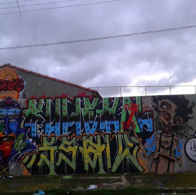 A colorful graffiti mural on a concrete wall reading 'WILL YOU BE BLUE FOR MY NAVAJA?' in large block letters, flanked by cartoon characters under an overcast sky.