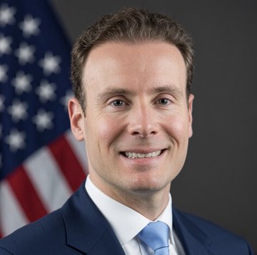 A professional headshot shows a smiling man in a dark suit and light blue tie, standing before an American flag, likely depicting CFTC Chair Mike Selig. The image accompanies a post about his pro-crypto regulatory stance and comments on the CLARITY Act driving financial innovation back to the U.S.