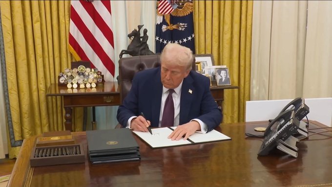 A man in a navy suit and red tie signs a document at the Resolute Desk in the Oval Office, surrounded by American flags and gold curtains. The image, captioned "AMERICA WILL LEAD ON CRYPTO GLOBALLY," visually reinforces the message by depicting a high-level official signing an executive order, symbolizing decisive U.S. action and leadership in the global crypto space.