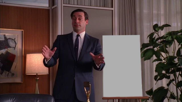 A man in a dark suit and tie stands in a mid-century room gesturing toward a blank canvas on an easel, next to a gold trophy.
